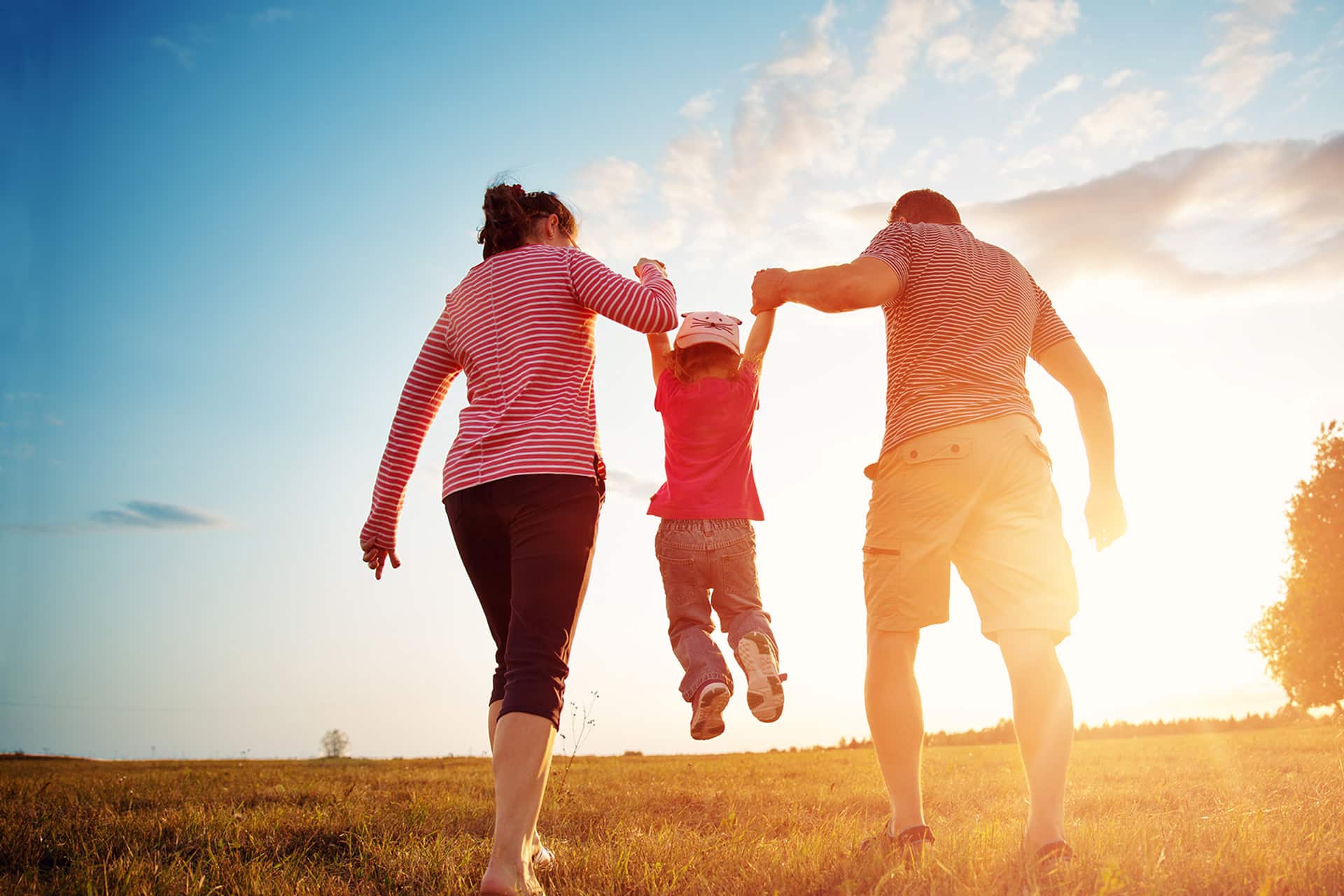 Young girl being lifted and swung in the air between mother and father. Walking through a field at sunset