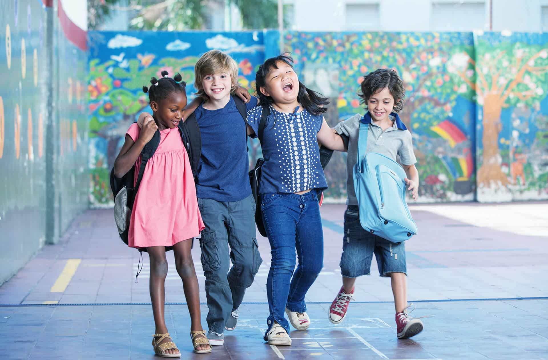 Smiling kids with their arms around each other and backpacks on walking out of school together.